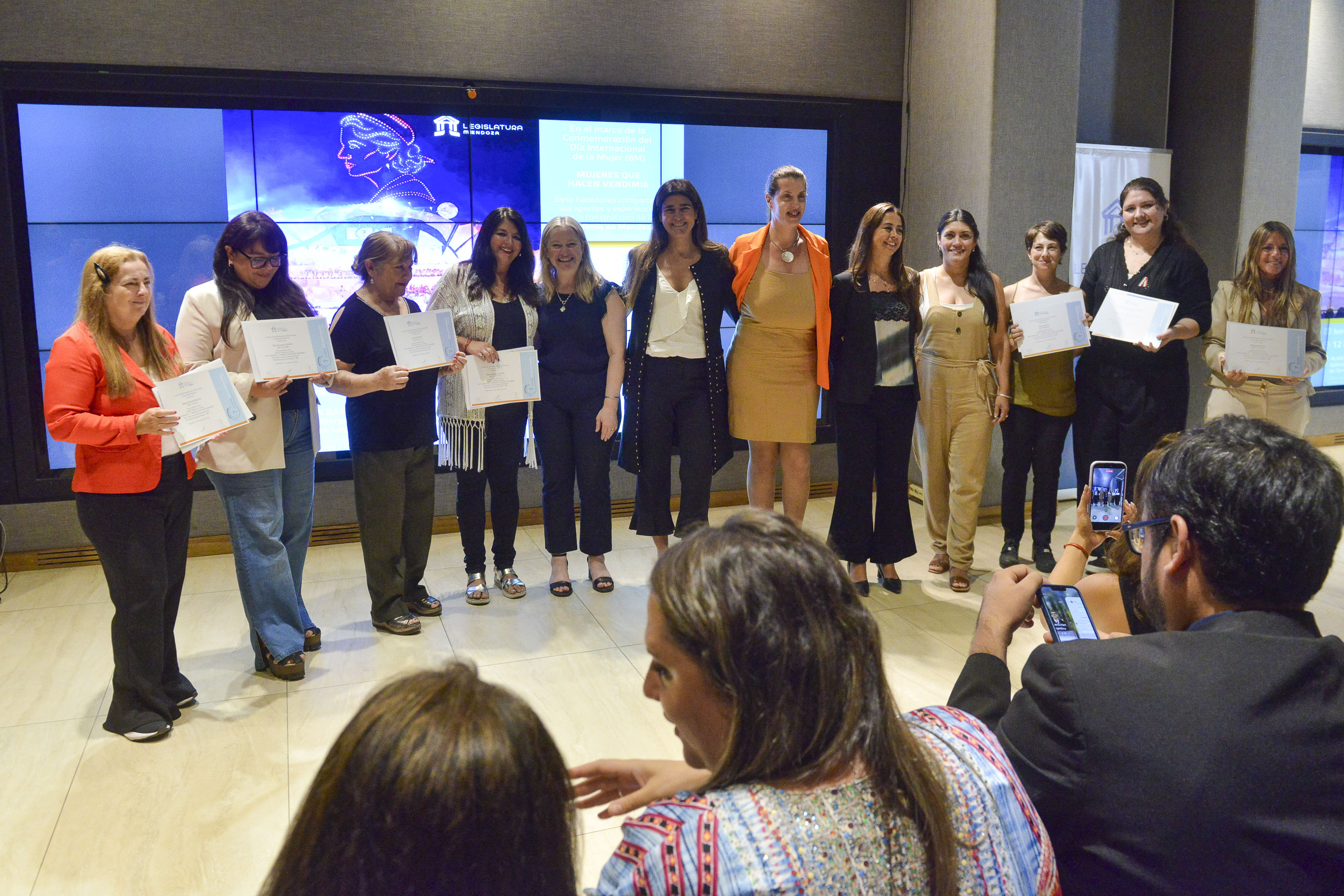 La actividad se realizó en el marco del Día Internacional de la Mujer y buscó visibilizar el aporte femenino en la industria vitivinícola y en la construcción cultural de la Vendimia. En la planta baja del edificio Margarita Malharro de Torres de la Legislatura de Mendoza, se llevó a cabo la charla “7 minutos, 7 mujeres que hacen Vendimia”, un encuentro que tuvo como objetivo visibilizar y destacar el aporte fundamental de las mujeres en la industria vitivinícola y en la cultura vendimial, en el marco de la conmemoración del Día Internacional de la Mujer. La actividad se desarrolló en el edificio ubicado en la Peatonal Sarmiento y reunió a mujeres vinculadas al mundo del vino y de la Vendimia desde distintos ámbitos. Cada una dispuso de siete minutos para compartir su experiencia, su historia y su aporte al sector, generando un espacio de reflexión, intercambio y reconocimiento. Participaron del encuentro Pamela Cuartara, directora de la Mujer, Género y Diversidad de la Suprema Corte de Justicia de Mendoza; Fernanda Urquiza, subdirectora de Género y Diversidad del Gobierno de Mendoza; Ramón Blecua, cónsul general de España en Mendoza; Luis Andrés García, cónsul adjunto del Consulado General del Perú en Mendoza; Nora Metz, decana de la Facultad de Ciencias Económicas y Jurídicas de la Universidad del Aconcagua; y Emiliana Lilloy, directora de Relaciones Institucionales y Escuela de Gobierno del Senado de Mendoza. Durante la jornada se destacó que la Vendimia, símbolo de la identidad mendocina, se construye a partir del trabajo de cientos de mujeres que participan en distintas etapas del proceso productivo y cultural, desde el viñedo hasta los escenarios artísticos y las actividades vinculadas a la promoción del vino. La identidad visual de la Vendimia La diseñadora gráfica Rita Carrizo relató su experiencia como parte del equipo que desarrolló la imagen gráfica del 90° aniversario de la Fiesta Nacional de la Vendimia. Explicó que el proyecto fue realizado junto a otros dos diseñadores mendocinos de la agencia Ilustre Pro y que implicó el desafío de sintetizar la historia y la identidad de la fiesta en una propuesta visual. Carrizo señaló que el equipo decidió tomar como referencia los primeros afiches vendimiales y colocar en el centro de la propuesta el rostro de una mujer mendocina, con el objetivo de destacar el trabajo humano detrás de la fiesta. También explicó que el diseño incorpora fotografías, ilustraciones y elementos simbólicos relacionados con la historia de la Vendimia y con distintos paisajes vitivinícolas de la provincia. La música en la fiesta central Por su parte, la directora orquestal Alicia María Pouzo, quien dirigió la música en las ediciones 2024 y 2026 de la Vendimia, compartió su recorrido dentro de la fiesta y reflexionó sobre el lugar de las mujeres en la dirección musical. Contó que su participación comenzó en 2023, cuando fue convocada para colaborar en la organización de los ensayos de la orquesta. Posteriormente, integró el equipo artístico en la edición 2024 y volvió a ser convocada este año. Durante su exposición destacó que la presencia de mujeres en la dirección orquestal todavía es reducida a nivel mundial, ya que entre el 8 y el 10 por ciento de las orquestas profesionales son dirigidas por mujeres. En ese sentido, valoró la oportunidad de que la Vendimia incorpore nuevos roles especializados dentro de su estructura musical. La danza como transmisión cultural La profesora Rosa Muñoz, directora del Ballet Lucerito, relató su trayectoria dedicada a la enseñanza del folklore y a la promoción de las tradiciones culturales. Explicó que hace más de 25 años fundó su grupo de danza con el objetivo de formar bailarines y transmitir identidad cultural a las nuevas generaciones. Además, mencionó la creación de una escuela de danza en Uspallata y su participación en diversas vendimias departamentales y en la fiesta central. Muñoz destacó que el folklore es un puente entre el pasado y el futuro, y que la cultura se construye colectivamente a través del aporte de artistas, músicos, bailarines y hacedores culturales. Una vida dedicada a la música cuyana La cantante y pianista Fabiana Cacace compartió su historia personal vinculada al mundo del vino y la música. Oriunda de San Rafael, recordó que pasó parte de su infancia en una bodega, donde su familia trabajaba, lo que marcó su relación con la cultura vitivinícola. Con más de treinta años de trayectoria en la música cuyana, ha participado en numerosos escenarios y celebraciones vendimiales. Durante su intervención interpretó un fragmento de una obra dedicada a la Vendimia, evocando el paisaje, el trabajo del viñedo y la identidad cultural de Mendoza. La organización de las reinas de la comunidad italiana La coordinadora de las reinas de la comunidad italiana, Karen Caballero Marini, relató su experiencia al frente de la organización de las representantes culturales vinculadas a la tradicional fiesta Pesca y Piazza. Contó que asumió la coordinación hace un año, luego de haber sido reina de una de las regiones italianas representadas en la celebración. Desde entonces, se propuso ampliar la participación de las reinas en distintos eventos culturales y protocolares. Según explicó, a partir de gestiones y contactos con municipios e instituciones logró que las representantes participaran en más de cincuenta actividades durante el último año. También destacó que su tarea implica coordinar a dieciséis jóvenes, acompañarlas en cada evento y resolver situaciones cotidianas vinculadas a la organización. Caballero Marini señaló que el rol requiere paciencia, compromiso y contención, y recordó con emoción el reconocimiento de las familias de las jóvenes al finalizar sus mandatos, quienes le agradecieron por el cuidado brindado durante las distintas actividades. El rol de las mujeres en la industria vitivinícola La licenciada en Administración de Empresas Marita Bressia, integrante de Bressia Winery, compartió su experiencia dentro de una bodega familiar fundada en 2001. Explicó que comenzó a trabajar junto a su padre en el área comercial cuando la empresa era muy pequeña y que, con el paso de los años, el emprendimiento fue creciendo hasta conformar un equipo de treinta personas. Destacó que actualmente la mitad del personal de la bodega está conformado por mujeres que se desempeñan en distintos sectores, desde el laboratorio y la recepción de uva hasta las áreas de turismo, hospitalidad y comercio exterior. Bressia señaló que en las últimas décadas se ha producido un cambio significativo en la industria vitivinícola, con una presencia cada vez mayor de mujeres ocupando distintos roles y participando activamente en el desarrollo del sector. La difusión del vino y la cultura vendimial La sommelier y periodista Gabriela Malizia, fundadora de Dionisias Wine Fair, relató su trayectoria en el periodismo y su vínculo con la Vendimia. Recordó que durante muchos años cubrió la fiesta para el diario UNO y que esa experiencia la llevó a profundizar su interés por el mundo del vino y por el rol de las mujeres dentro de la industria. En ese contexto surgió la idea de crear una feria de vinos protagonizada exclusivamente por mujeres enólogas, ingenieras agrónomas y sommeliers. La iniciativa comenzó con un pequeño encuentro y con el tiempo fue creciendo hasta consolidarse dentro del calendario cultural de la provincia. Malizia explicó que el objetivo de Dionisias Wine Fair es dar visibilidad al trabajo de las mujeres del sector y acercar la cultura del vino a nuevos públicos. Reconocimiento y cierre de la jornada Al finalizar la actividad, Emiliana Lilloy junto a Fernanda Urquiza, Pamela Cuartara y Nuria Olivera, directora de la Secretaría Legal y Técnica del Senado de Mendoza, entregaron reconocimientos a las expositoras en agradecimiento por su trayectoria y por el aporte que realizan a la cultura vendimial. El cierre estuvo a cargo de la vicegobernadora Hebe Casado, quien destacó el rol de las mujeres en el desarrollo de la provincia. Señaló que la intención del encuentro fue visibilizar a mujeres que, desde distintos ámbitos, impulsan el crecimiento y el progreso de Mendoza. La vicegobernadora remarcó que en cada actividad vinculada a la Vendimia hay mujeres que trabajan y sostienen distintos espacios, y agradeció a las expositoras por compartir sus experiencias y por representar a tantas mendocinas que también contribuyen a la construcción de la identidad provincial. La jornada concluyó con un ágape destinado a promover el encuentro y el intercambio entre los presentes, acompañado por vinos de la Bodega Sueños de Revolución.